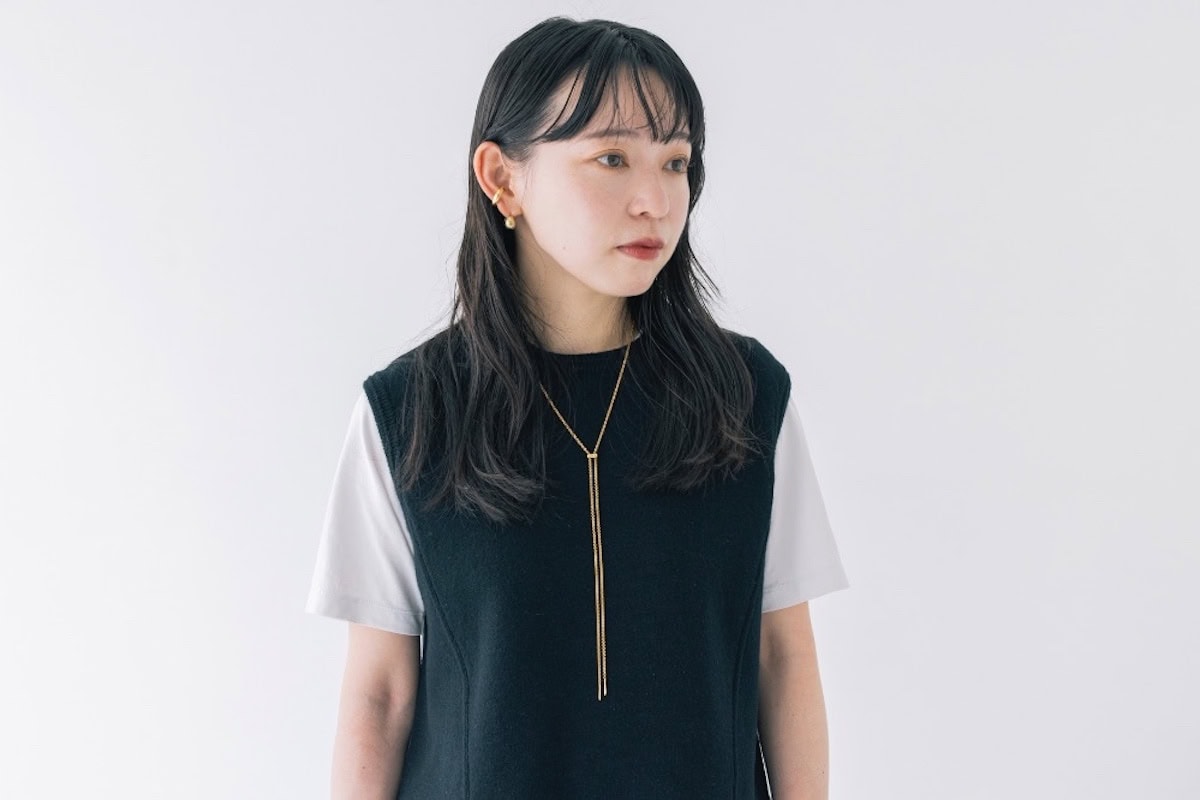 淡いグレーのTシャツにティッカの黒いメリノウールベストを重ね、プロウのゴールドネックレスと耳もとのアクセサリーを合わせた上半身